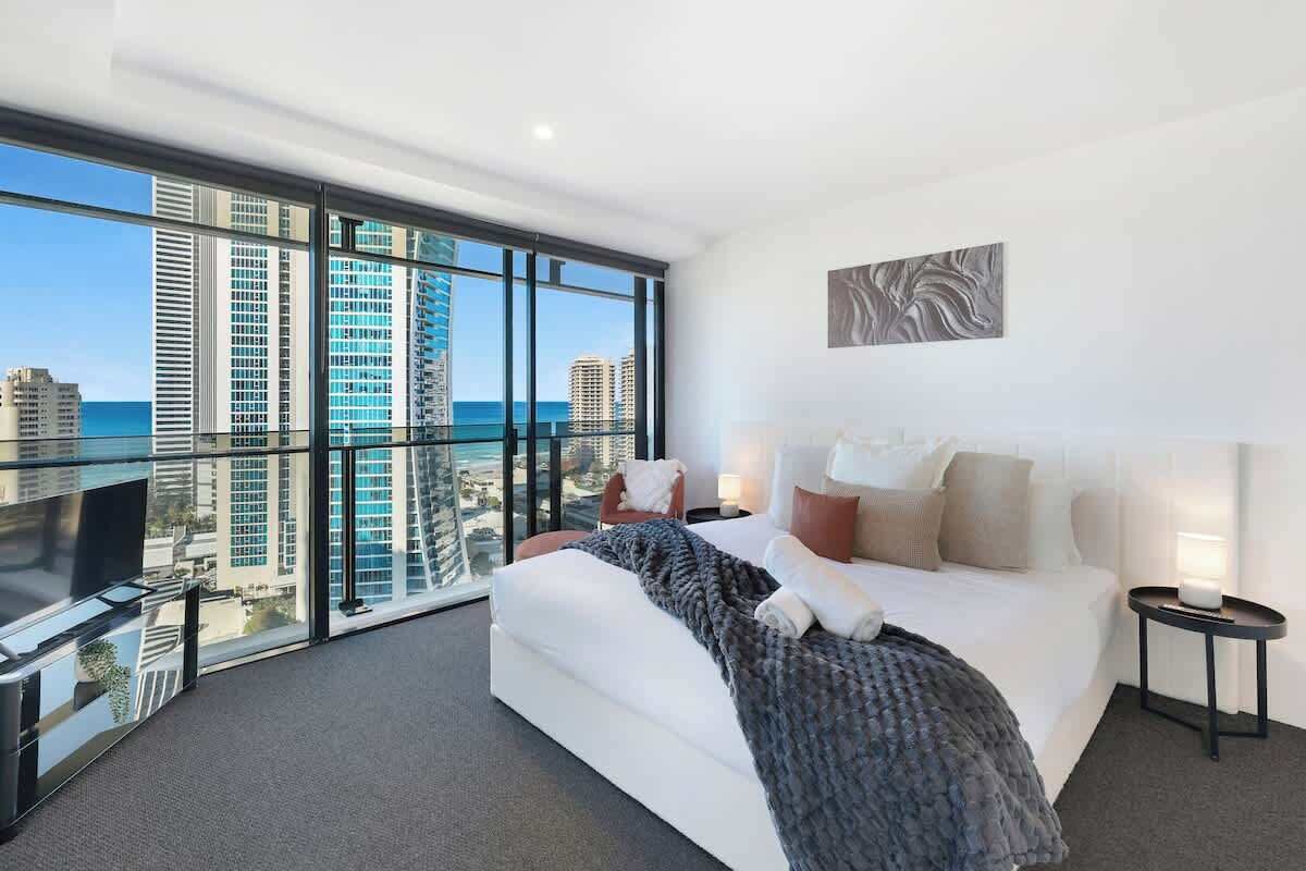 A modern hotel room with a large bed, a view of the cityscape, and a balcony with a glass railing.
