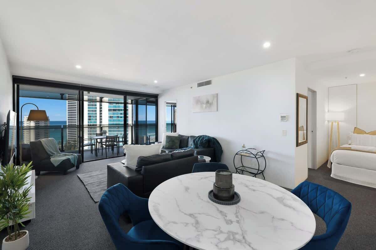 A modern living room with a round marble table, blue chairs, a sofa, and a balcony with a view of the city and ocean.