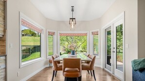 Un comedor con una mesa redonda, seis sillas y ventanas amplias que ofrecen vistas a un paisaje verde.
