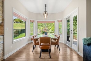 A dining area with a round table, six chairs, and large windows offering a view of a green landscape.