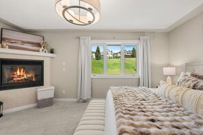 A bedroom with a fireplace, a large window with a view of a green landscape, a bed with a fluffy blanket, and a modern chandelier.