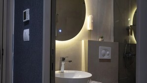 A modern bathroom with a white sink, a mirror, a wall-mounted light, and a shower. - NAM HOTEL (Prizren)