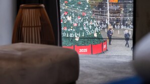 A window view of a Christmas tree with decorations, a sign reading '2025', and a glimpse of buildings outside. - NAM HOTEL (Prizren)