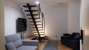 A modern living room with a grey sofa, a dark sofa, a wooden staircase, and a flat-screen TV mounted on the wall. - NAM HOTEL (Prizren)