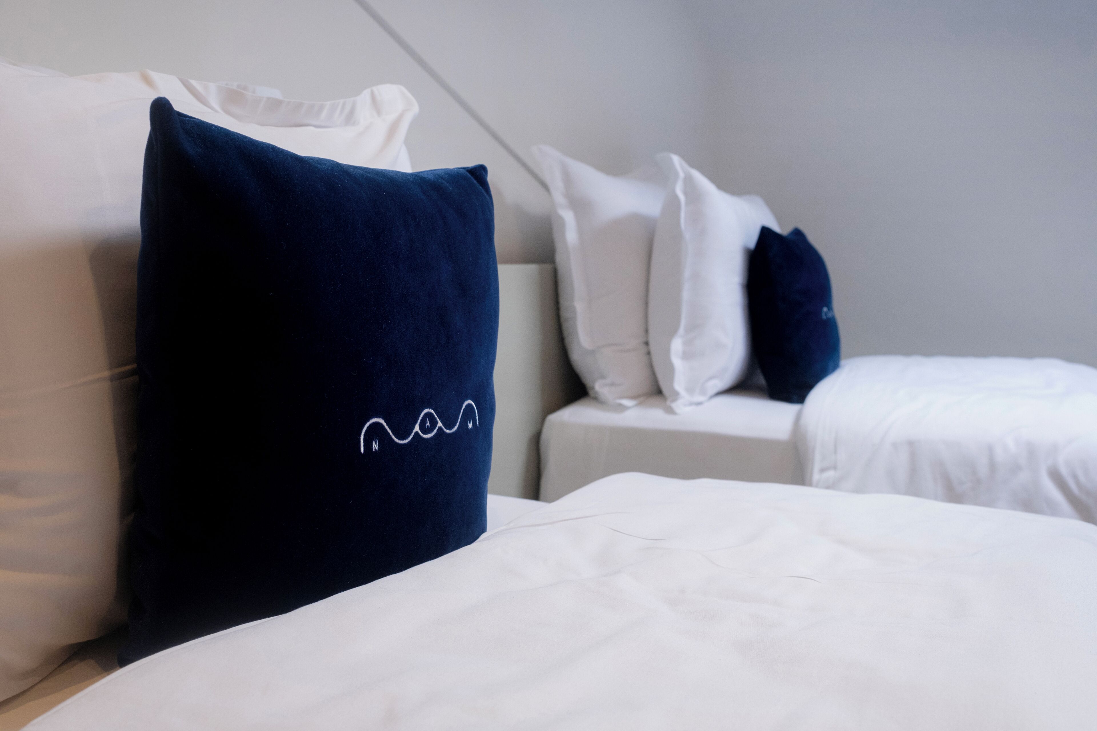 Una habitación de hotel con una cama, dos mesitas de noche blancas y un cojín azul oscuro con diseño blanco.
