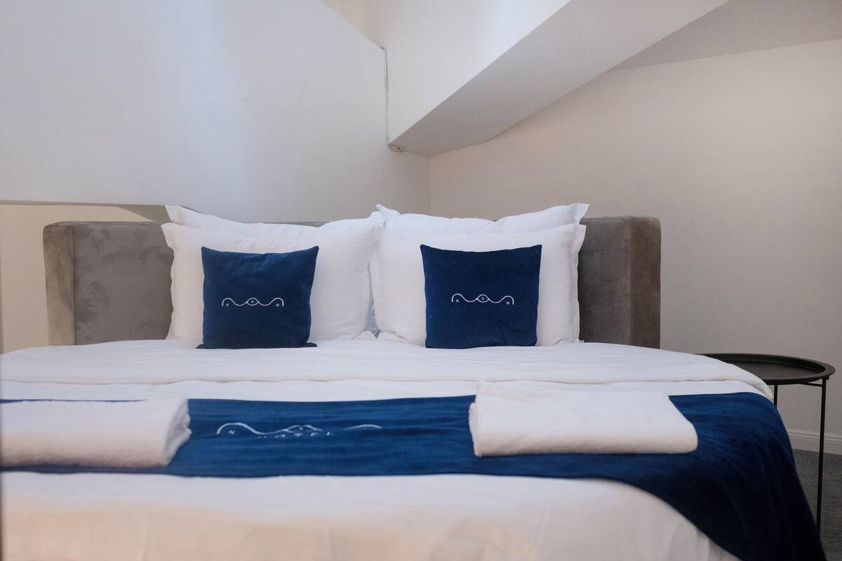 Una cama bien hecha con ropa de cama blanca y azul, un cabecero tapizado en gris y una pequeña mesa redonda al lado.