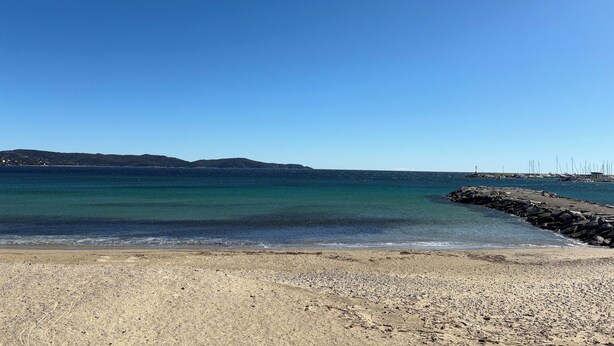 En strand med klart vatten, en stenig kust och kullar i fjärran.