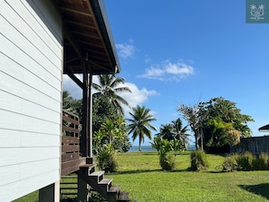 A wooden building with a covered porch and a view of a grassy area with palm trees and a body of water.