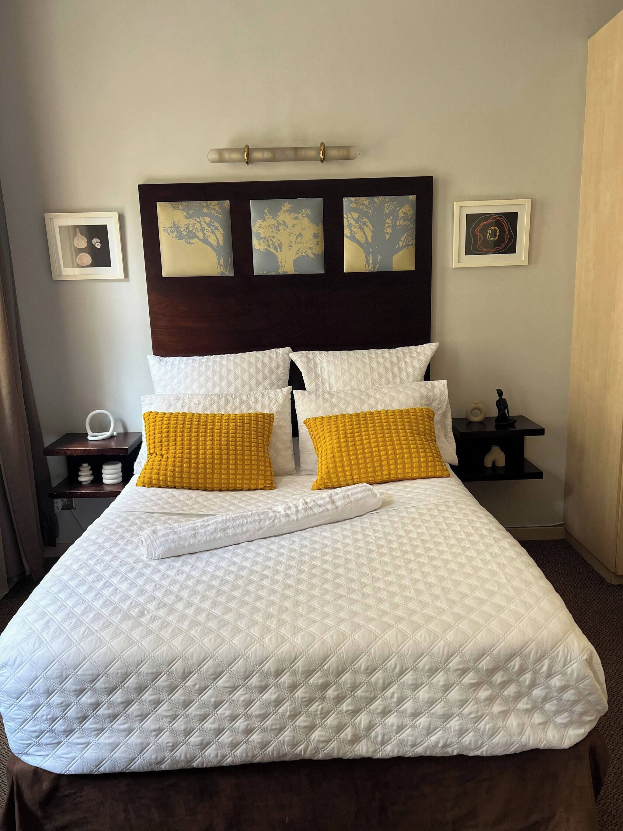 A neatly made bed with a quilted white bedspread, two pillows with a yellow textured cover, and a wooden headboard with three framed artworks.