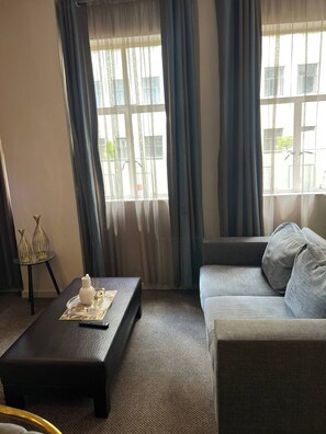 Una sala de estar con un sofá, una mesa de centro con adornos, una mesita auxiliar con jarrones y ventanas amplias con cortinas.