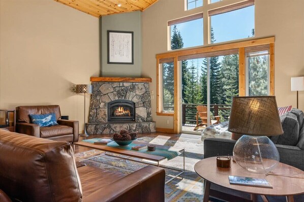 A cozy living room with a stone fireplace, leather furniture, and large windows offering a view of trees. - Ski Slope Views Tahoe Donner Retreat (Truckee)