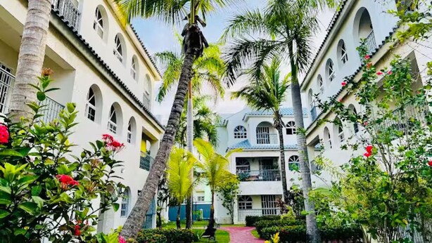 A courtyard with a central pathway flanked by buildings with arched windows and balconies, surrounded by palm trees and tropical plants. - Cozy 2BR/2Bth walk 2 the beach. Pool view & Wi-Fi (Punta Cana)