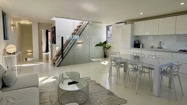 A modern kitchen with white cabinets, a dining area with white chairs, and a staircase leading to another level. - White Loft - 6 personnes - toit-terrasse avec vue panoramique (Donzenac)
