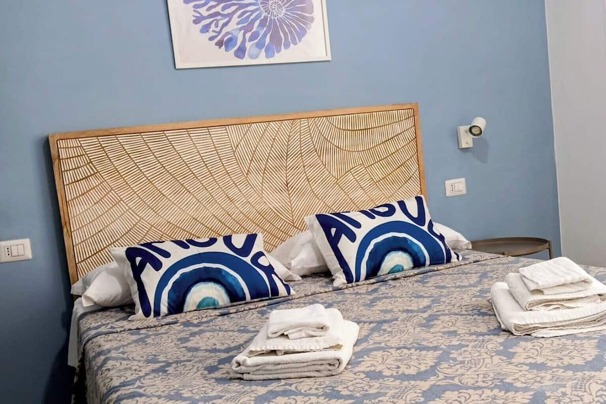 Siististi pedattu sänky, jossa on kuviollinen vuodevaatesetti, kaksi sinivalkoista koristetyynyä ja seinällä, sängyn yläpuolella, oleva kehystetty taideteos.