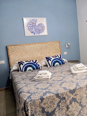 Ein ordentlich bezogenes Bett, Bettwäsche mit Muster, zwei Kissen mit blau-weißem Design und ein gerahmtes Kunstwerk an der Wand darüber.