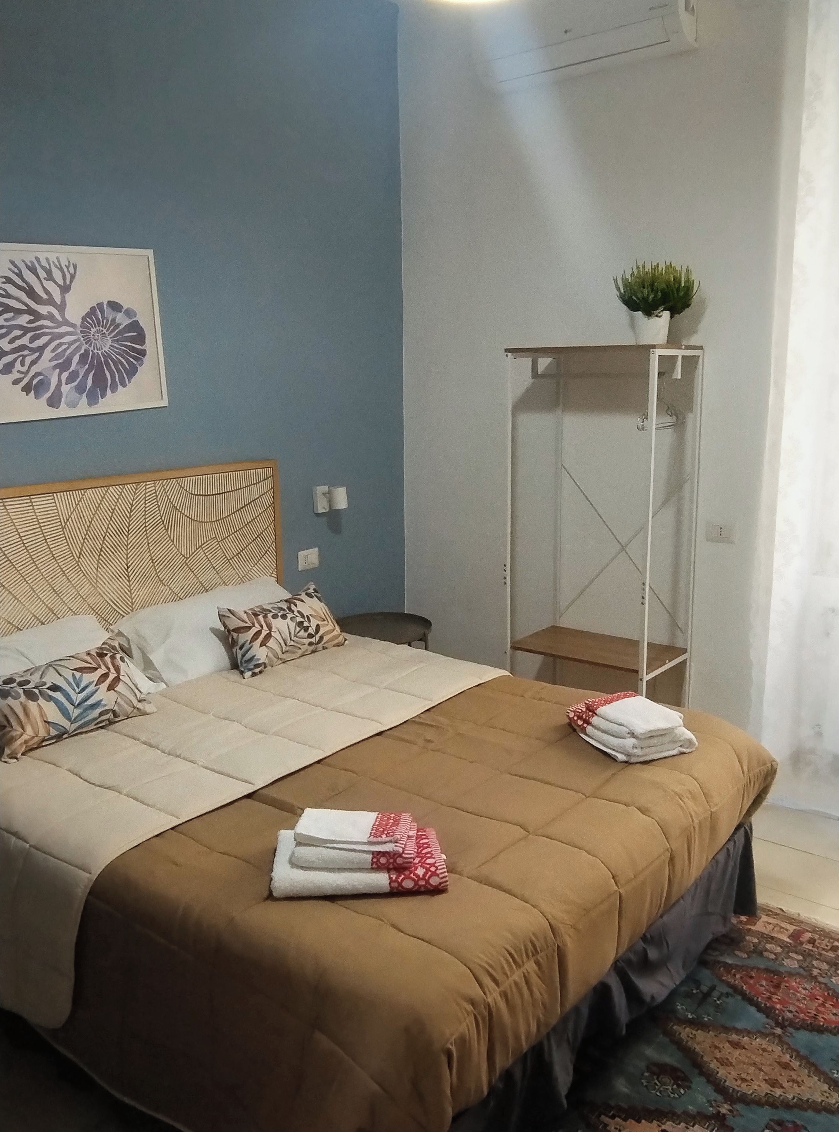 Ein ordentlich bezogenes Bett mit einer braunen Tagesdecke, zwei Kissen und einem gefalteten Handtuch an der Seite. An einer an der Wand befestigten Ablage steht eine Pflanze, und über dem Bett hängt ein gerahmtes Kunstwerk.
