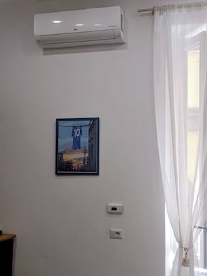 Huoneessa on seinään asennettu ilmastointilaite, seinällä on kehystetty taulu ja ikkunassa on ohuet verhot.