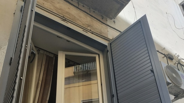 Ein Fenster mit halb geöffnetem Rollo, einer sichtbaren Klimaanlage und einem Balkongeländer.