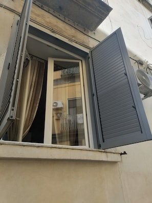 Una ventana con una persiana entreabierta, un equipo de aire acondicionado a la vista y una barandilla de balcón.