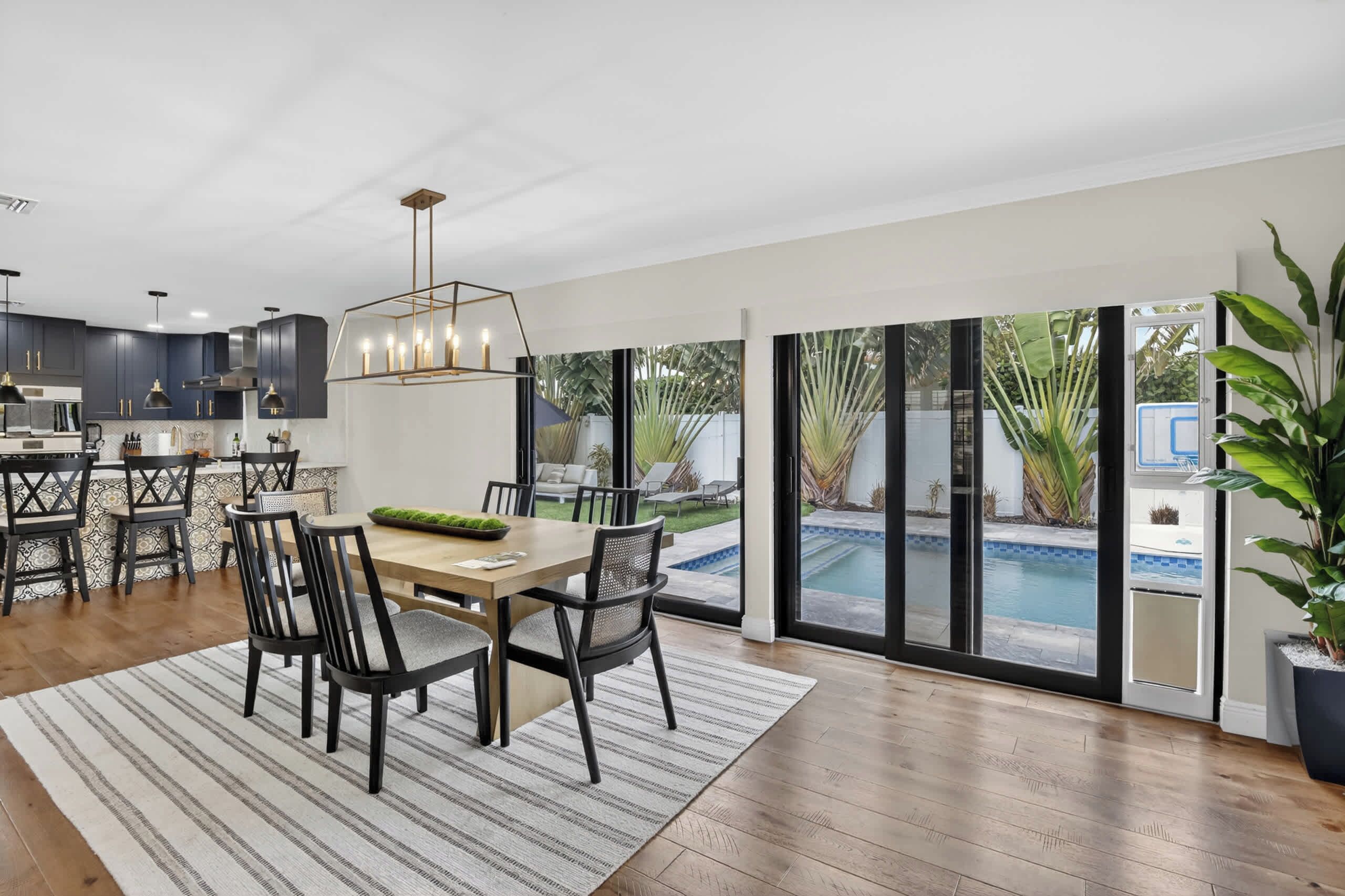 A modern kitchen with dark cabinetry, a dining area with a wooden table and chairs, and large sliding glass doors leading to a pool.