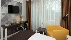 Hotellihuone, jossa on sänky, työpöytä tuolilla, seinälle asennettu televisio, pieni pöytä lampun kanssa ja keltainen nojatuoli.