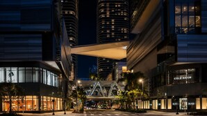 Une rue moderne d’une ville, de nuit, avec des bâtiments illuminés, un pont piétonnier et des devantures de magasins, dont un magasin Cole Haan.