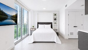 Une chambre moderne avec un grand lit, un téléviseur fixé au mur et une vue sur les immeubles et les arbres.