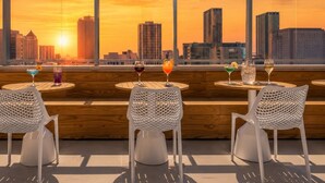 Un bar situé sur un toit, avec des chaises et des tables en treillis blanc, offrant une vue imprenable sur la ligne d’horizon de la ville au coucher du soleil.