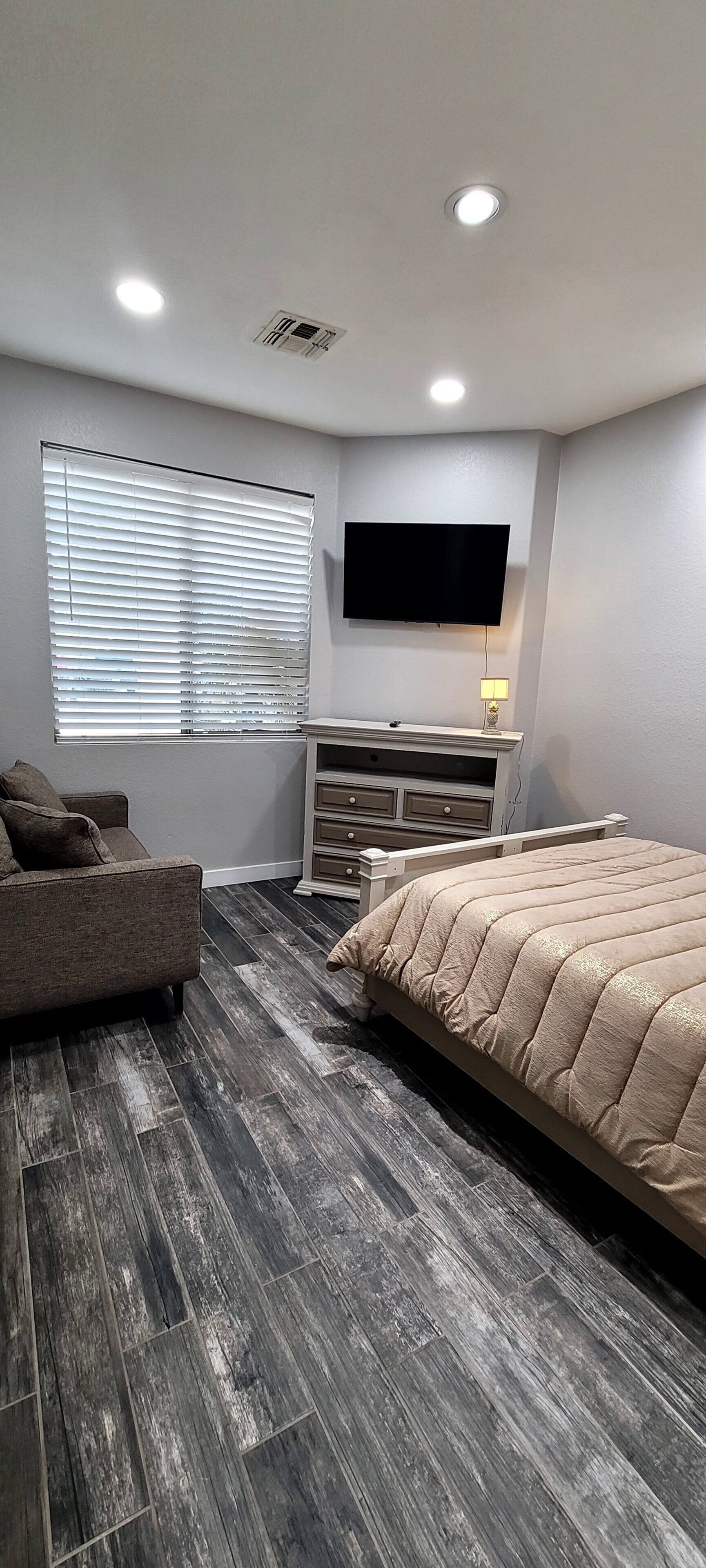 Quarto com cama, televisão na parede, janela com persianas, mesa de cabeceira com abajur e cômoda.