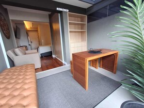 Quarto compacto com cama, uma pequena mesa de madeira, um sofá e porta deslizante para um banheiro.
