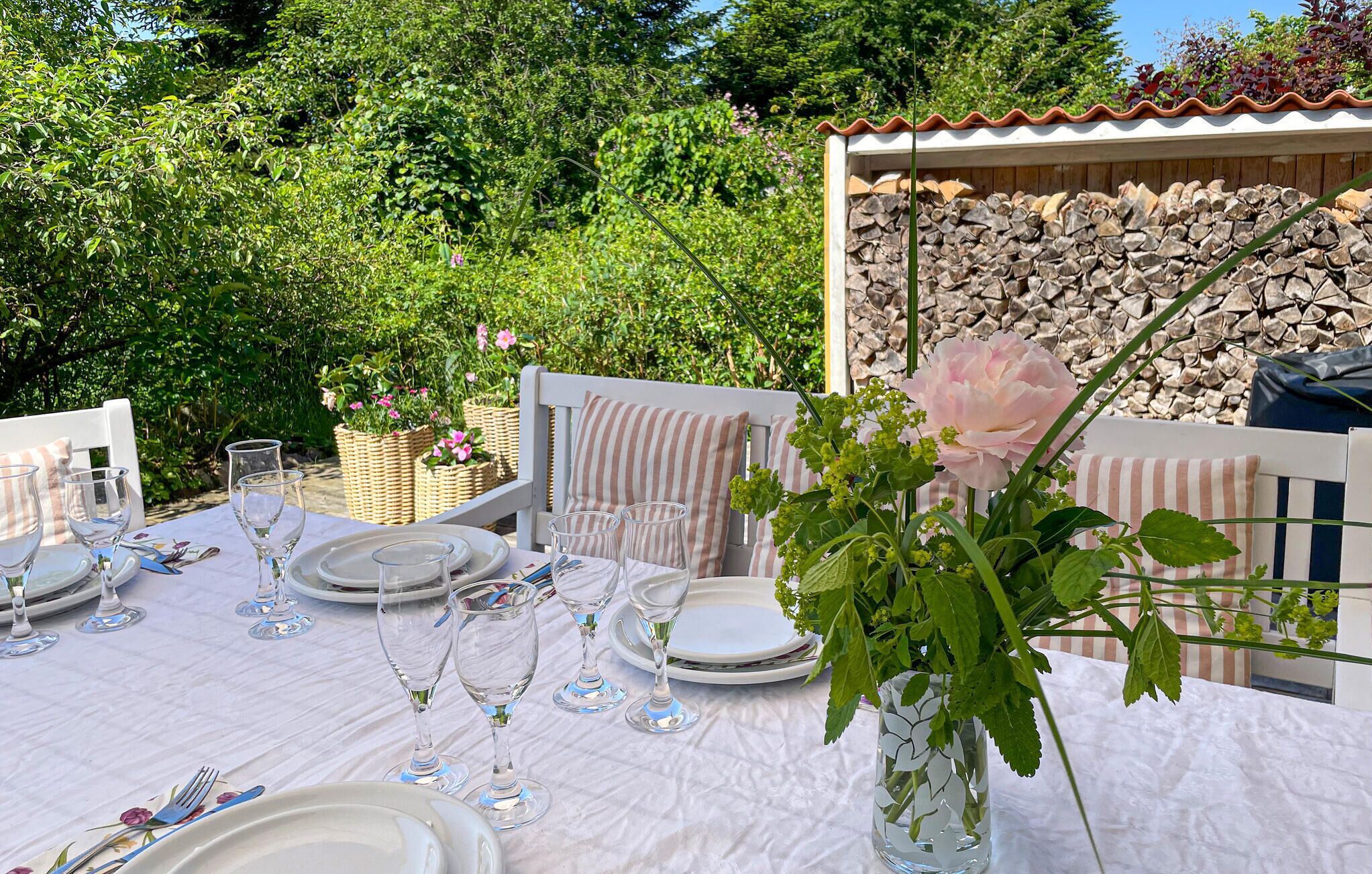 Un espace de restauration en plein air avec une table recouverte d’une nappe blanche, mise avec des assiettes, des verres et des couverts. On y voit des paniers en osier remplis de fleurs et, en arrière-plan, un mur de pierre.