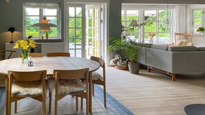 Un área de comedor con una mesa redonda, sillas y un jarrón de flores. Hay un sofá y una ventana con vistas a zonas verdes.