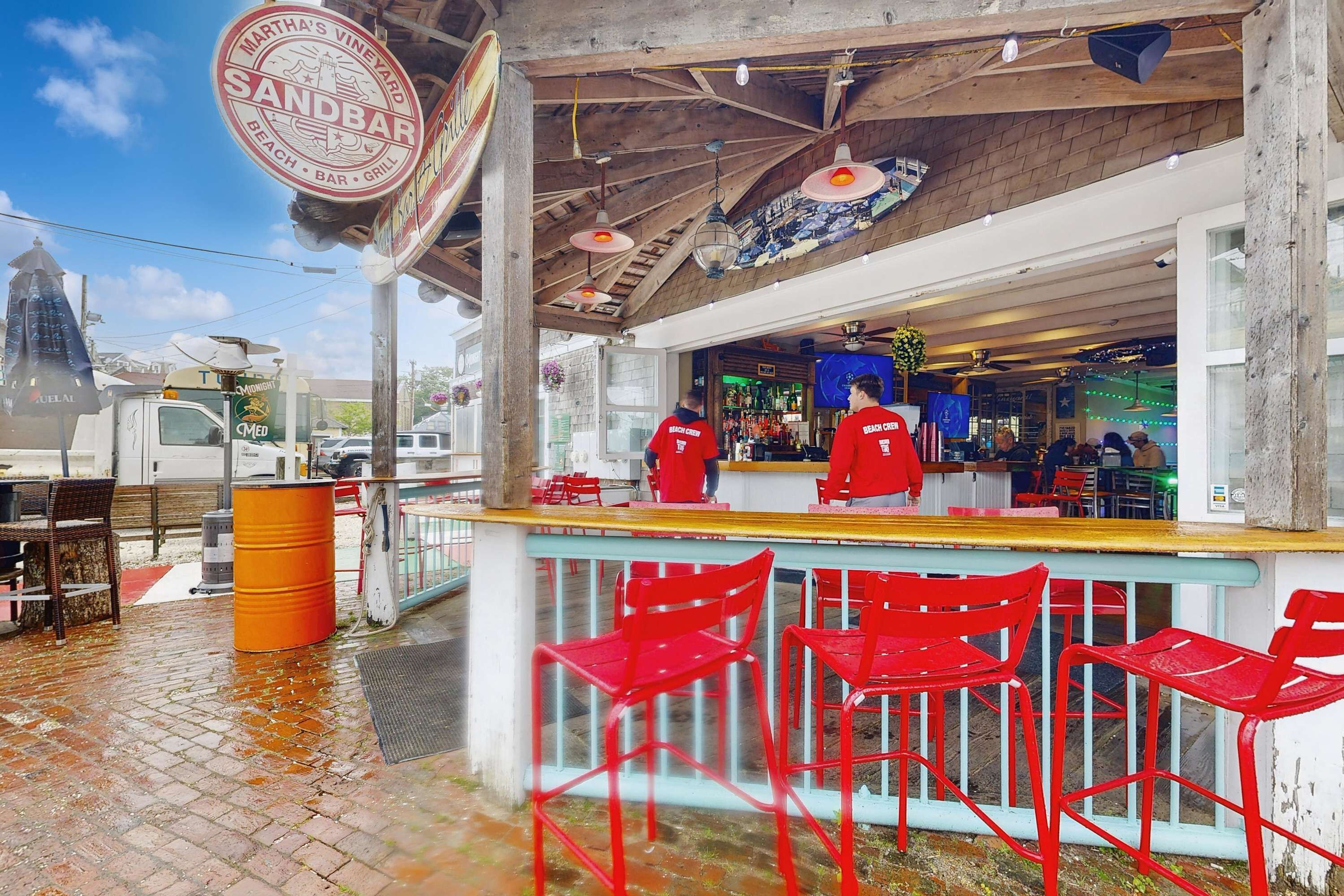 A beach bar with a wooden ceiling, red chairs, and a counter with staff in red uniforms.