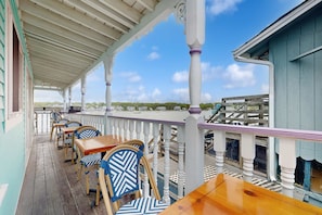 A covered outdoor seating area with wooden tables and chairs, a white railing, and a view of the surrounding buildings.