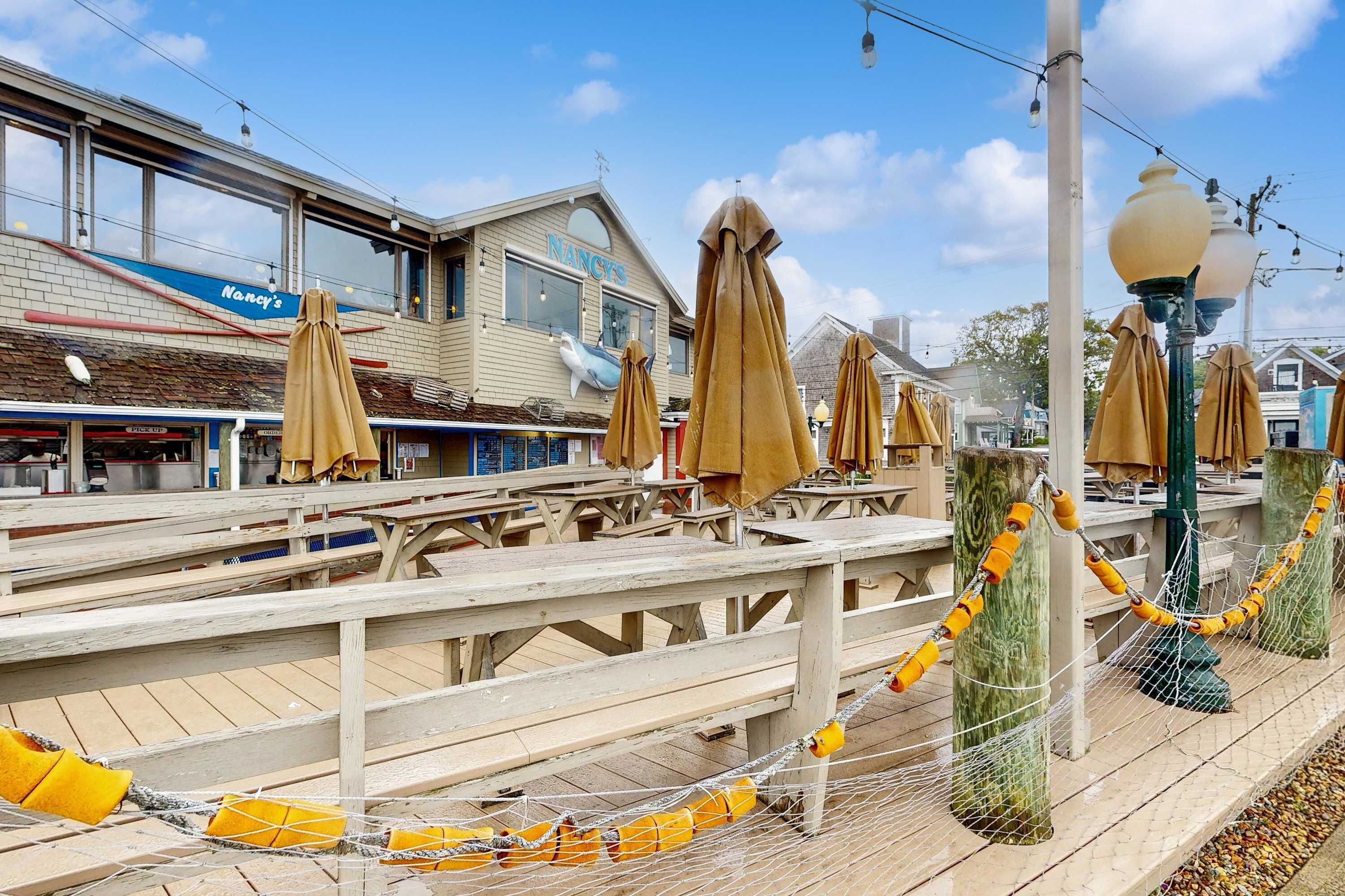 A wooden pier with tables and chairs, a building with a sign reading 'Nancy's,' and a street lamp.