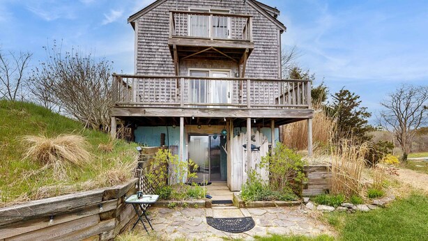 A two-story wooden house with a balcony, a stone pathway leading to the entrance, and a small garden area.