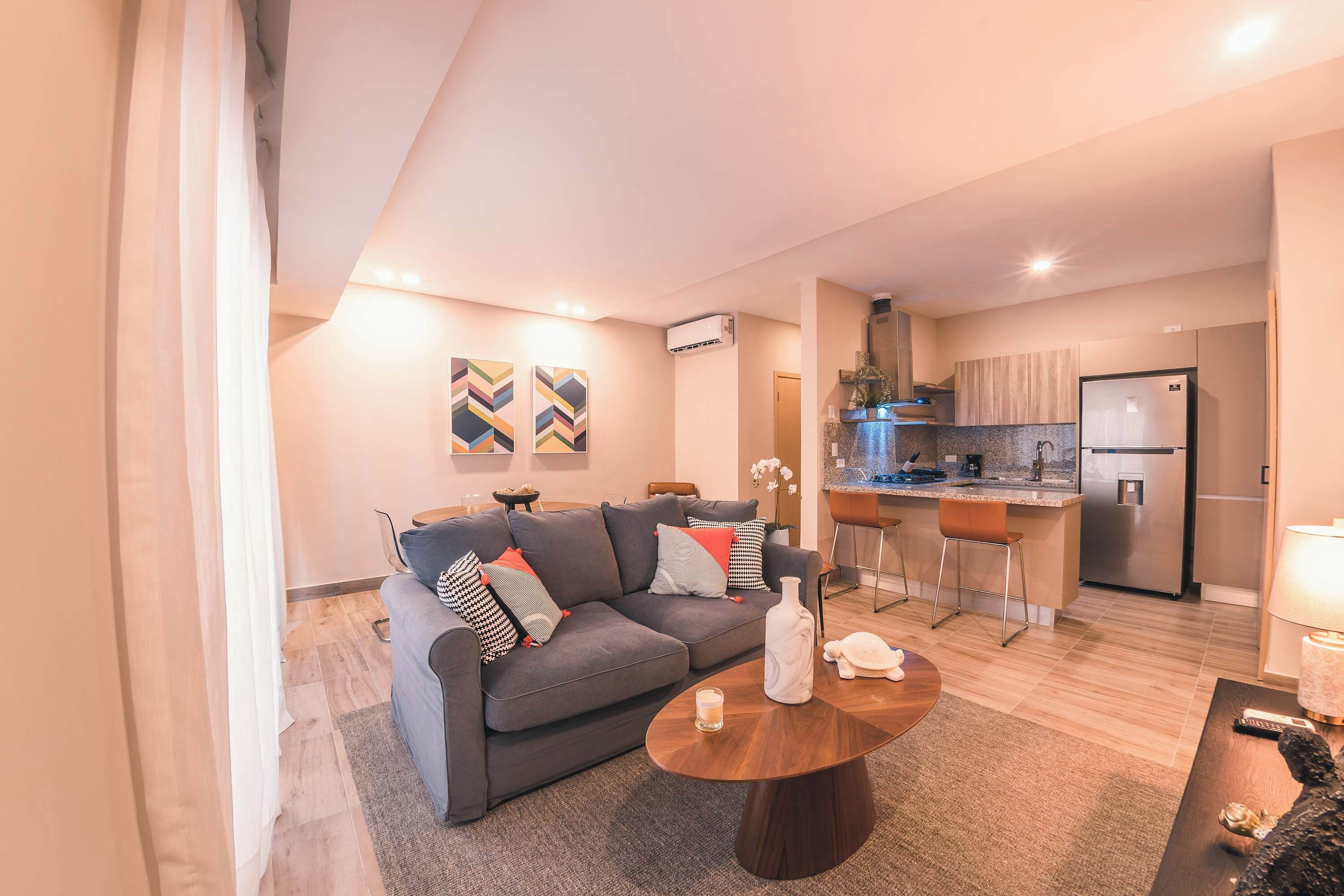 A modern living room with a grey sofa, a coffee table, and a kitchen area with a fridge and bar stools.