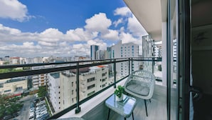 A balcony with a view of a cityscape, a wicker chair, a small table with a plant, and a glass partition. - AEA- Calle Victor Garrido Puello (Santo Domingo,)