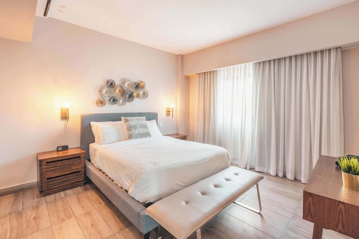 Ein modernes Schlafzimmer mit einem großen Bett, Nachttischlampen, einer Bank und Blick ins Freie durch Vorhänge.