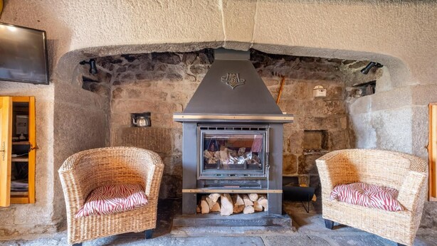 A stone fireplace with a modern wood-burning stove, two wicker chairs with striped cushions, and a stone wall. - Les Fleurs de l'Aubrac 4/6 pers (SAINTE-COLOMBE-DE-PEYRE)