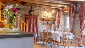 A rustic interior with a stone wall, wooden beams, a dining table set for four, and a vase of flowers on a counter. - Les Fleurs de l'Aubrac 4/6 pers (SAINTE-COLOMBE-DE-PEYRE)