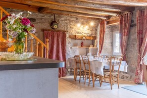 Ein rustikaler Innenraum mit einer Steinwand, Holzbalken, einem Esstisch für vier Personen und einem Blumenstrauß auf einer Anrichte.