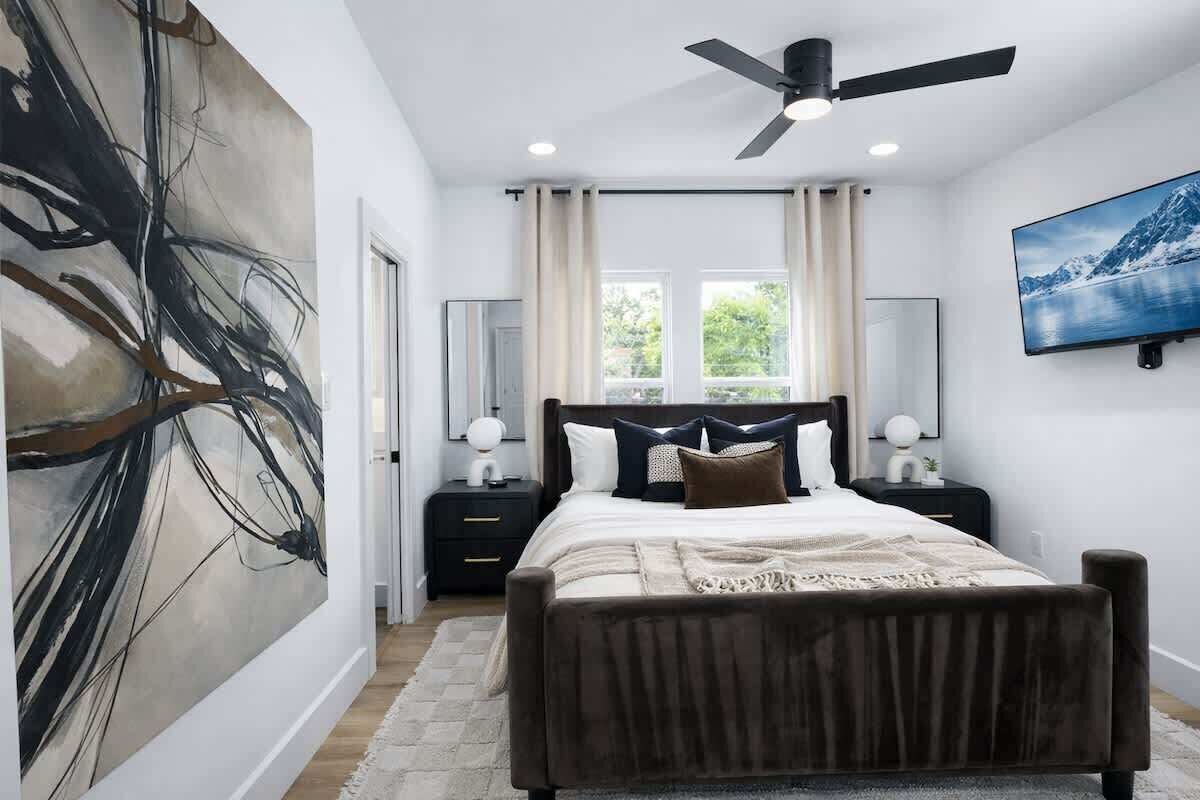 A bedroom with a bed, a ceiling fan, a large abstract painting, a window with curtains, and a TV on the wall.