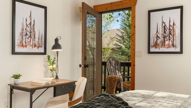 A bedroom with a bed, a desk, a chair, two framed artworks, and a view through an open door.
