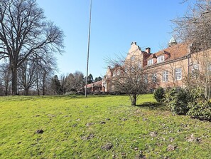 Ein großes Backsteingebäude mit mehreren Schornsteinen, einer grünen Wiese und kahl stehenden Bäumen.