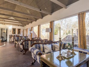 Ein geräumiges Zimmer mit hoher Decke, Holzbalken, einem großen Fenster mit Vorhängen, einer Sofagarnitur und einem Tisch mit Kerze und Vase.