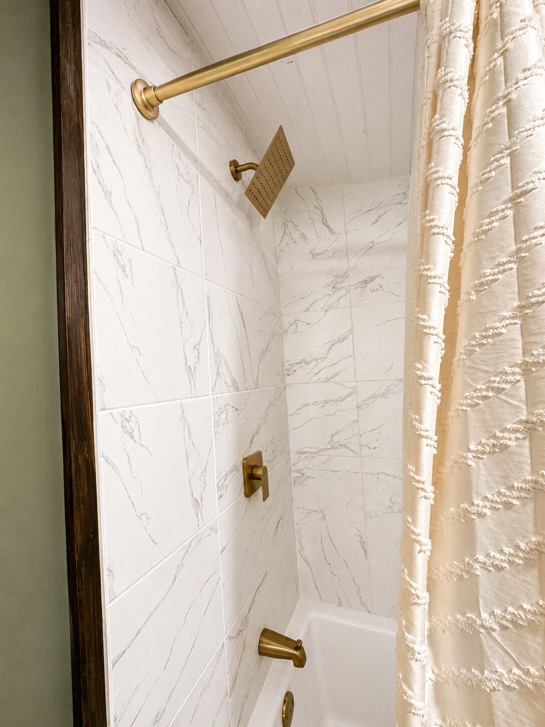 A bathroom with white marble tiles, a gold showerhead, a gold faucet, and a beige shower curtain.