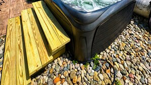 A hot tub with a wooden bench, surrounded by pebbles and greenery.