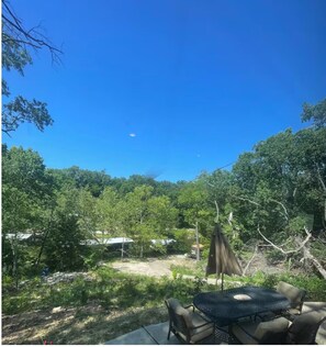 Eine Terrasse mit rundem Tisch und Stühlen, umgeben von Bäumen und blauem Himmel.