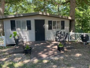 A small, single-story building with a front door, windows, a deck, outdoor furniture, and a grill. - Quaint Cabin at The Lake! (Camdenton)